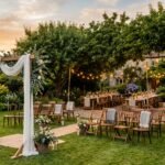A small backyard wedding at golden hour with a draped arch, lawn seating, and a long table under string lights