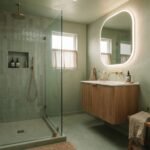 Small sage-green color-drenched bathroom with curbless glass-panel shower, floating wood vanity and oversized backlit mirror — small bathroom trends 2026