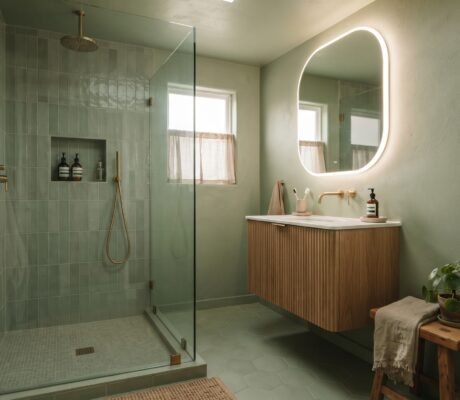 Small sage-green color-drenched bathroom with curbless glass-panel shower, floating wood vanity and oversized backlit mirror — small bathroom trends 2026