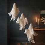 Three cheesecloth ghosts hanging at staggered heights against a charcoal wall, lit by warm candlelight