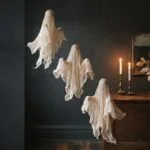 Three cheesecloth ghosts hanging at staggered heights against a charcoal wall, lit by warm candlelight